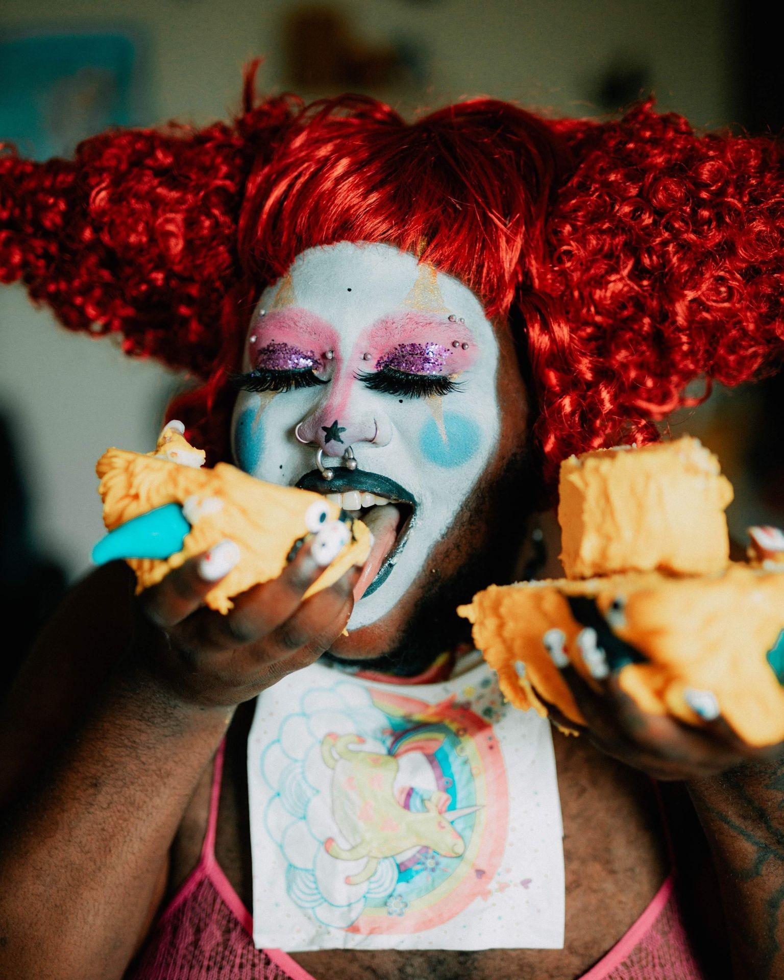 Person in colorful makeup and red wig eating cake, wearing a unicorn-themed shirt.