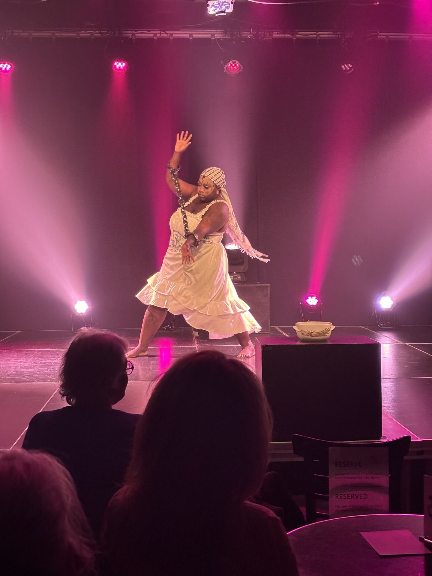 Performer in white dress dancing on stage with dramatic lighting and audience silhouettes.