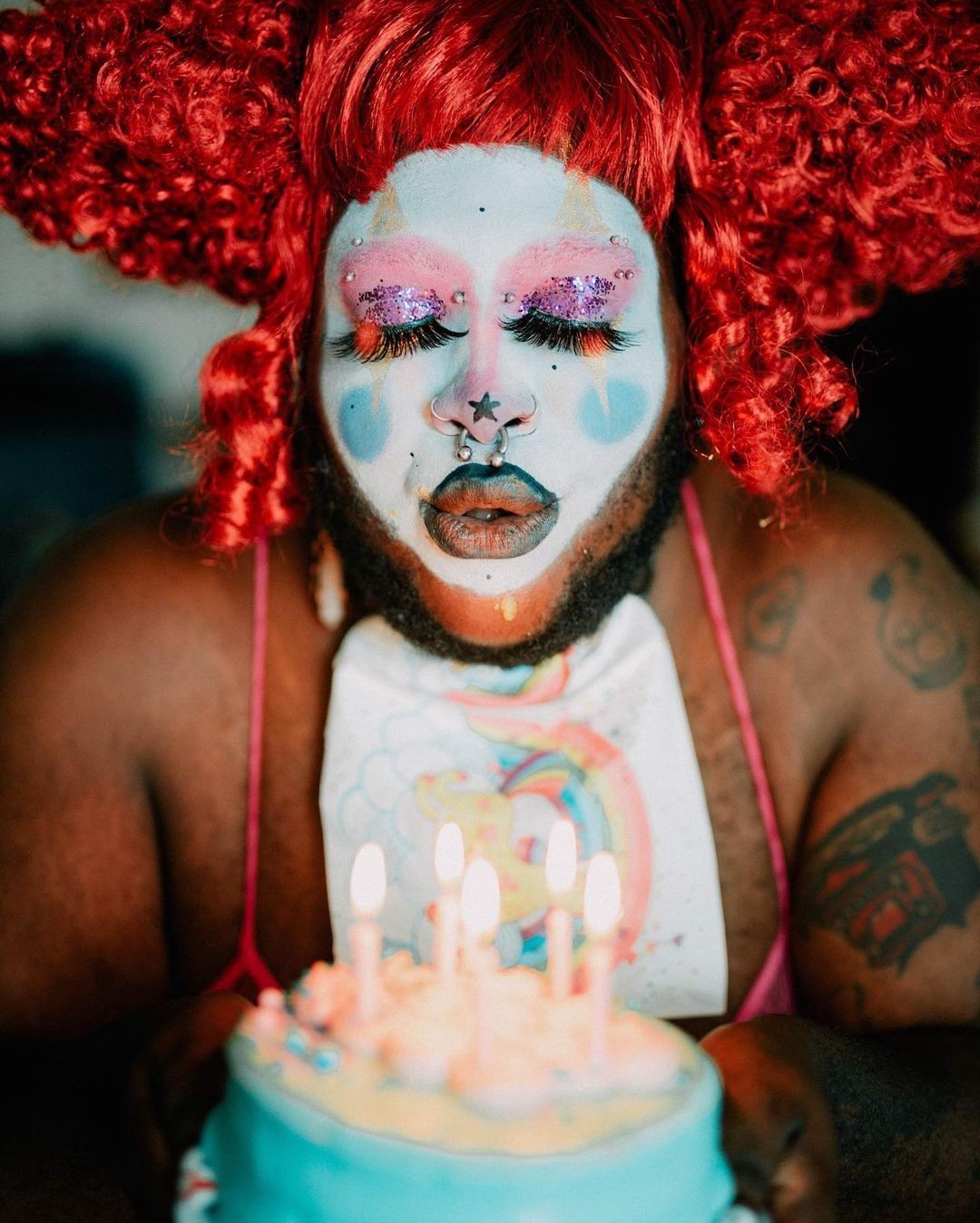 Person with colorful makeup and red wig blowing out candles on a birthday cake.