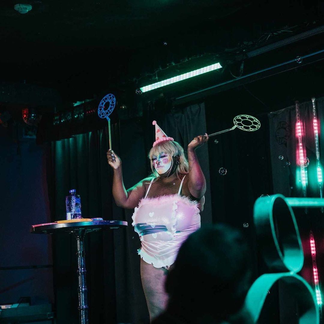 Performer in colorful costume and party hat on stage with bubble wands under vibrant lighting.