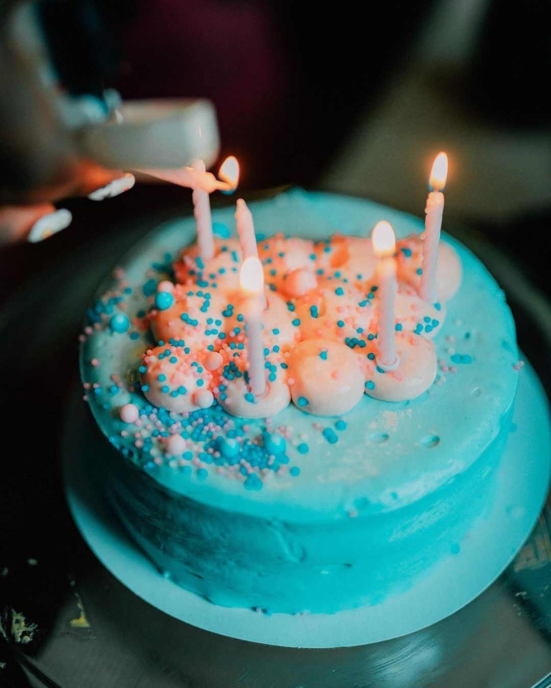 Birthday cake with blue frosting and five lit candles, decorated with pink and blue sprinkles.