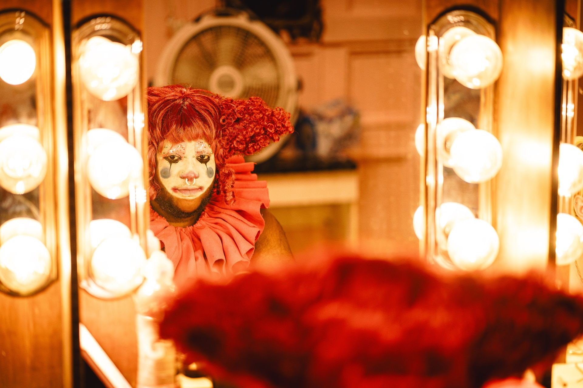 Clown with colorful makeup and curly red wig reflected in a mirror with bright lights.